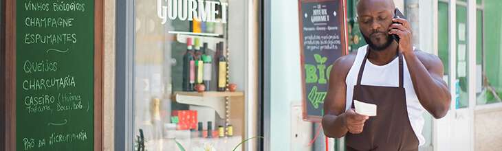 Dark-skinned man in a trendy grocer apron uses mobile phone outside a boutique gourmet shop